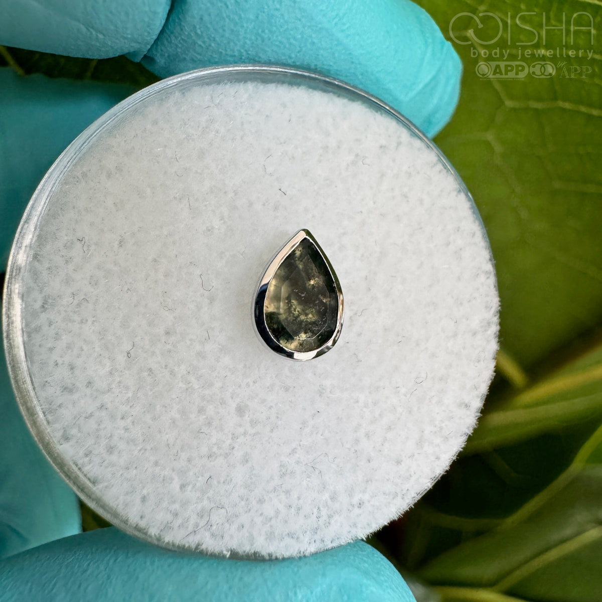 Teardrop-shaped gemstone in a white gold setting on a white cushion with green leaves in the background
