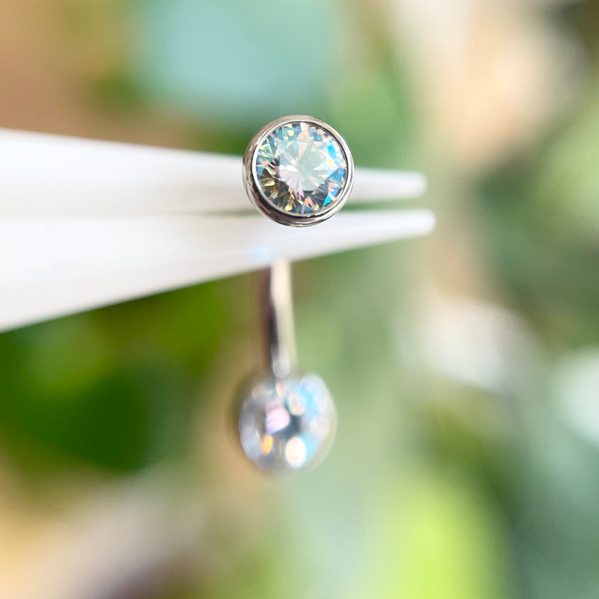 Silver looking belly bar with a iridescent gemstone on a white stand against a blurred green background