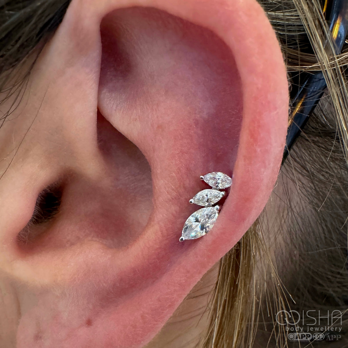 Three pear-shaped marquis-cut cluster earrings on a person's ear known as an auricle helix piercing with 'Isha Jewellery' and 'association of professional piercers member' branding.