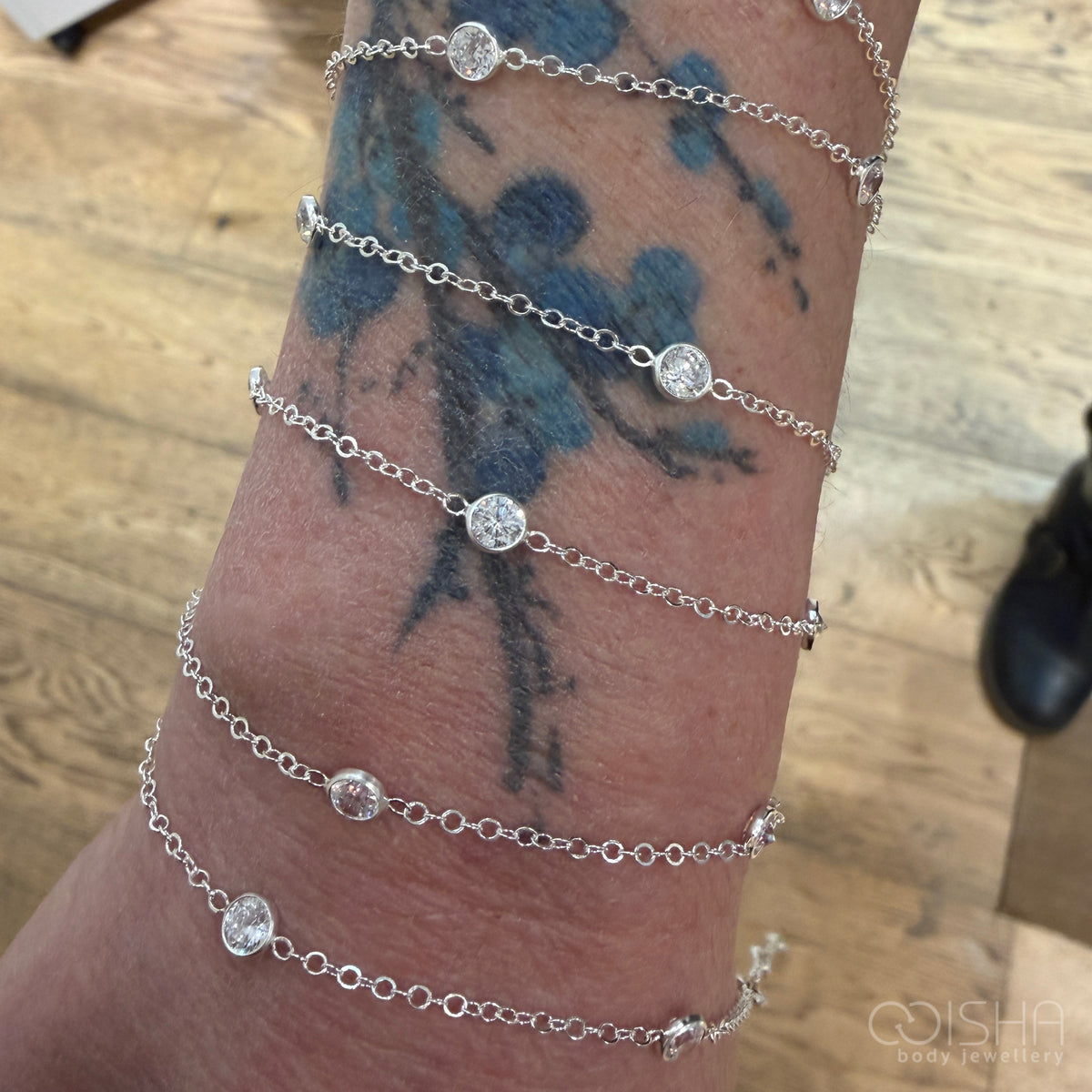 Silver chain bracelet with clear gemstones on a wrist with tattoos, against a wooden surface.
